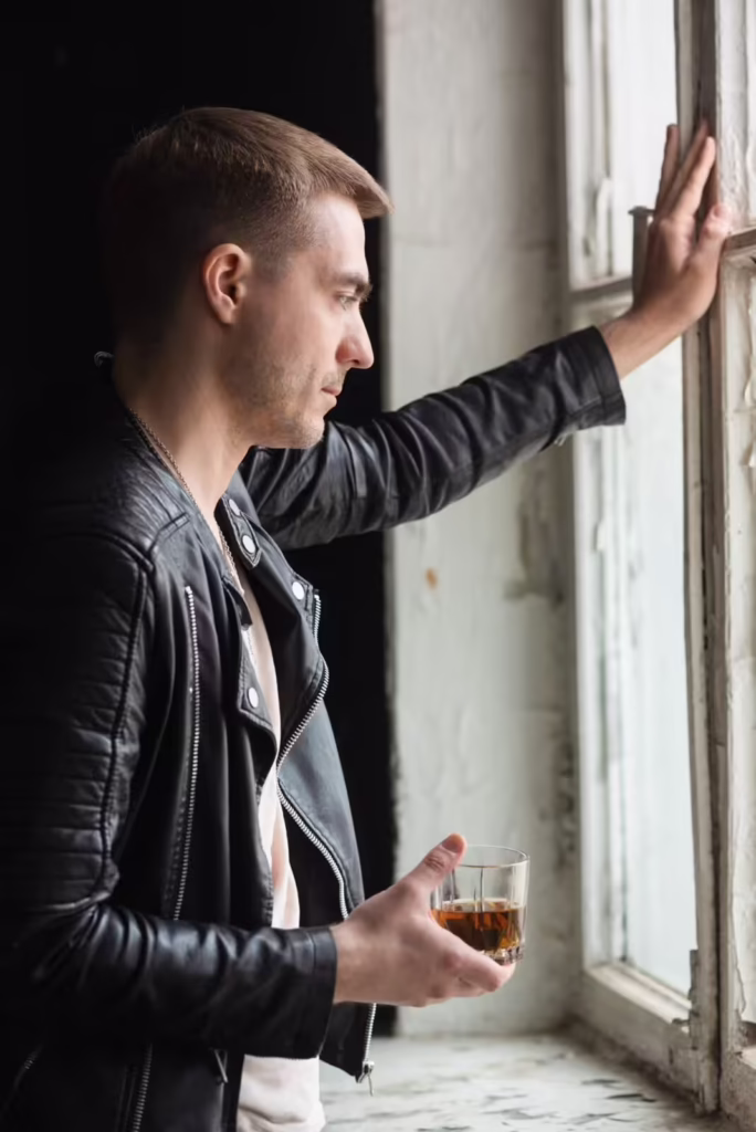 A man holding an alcoholic beverage and sadly looking out a window